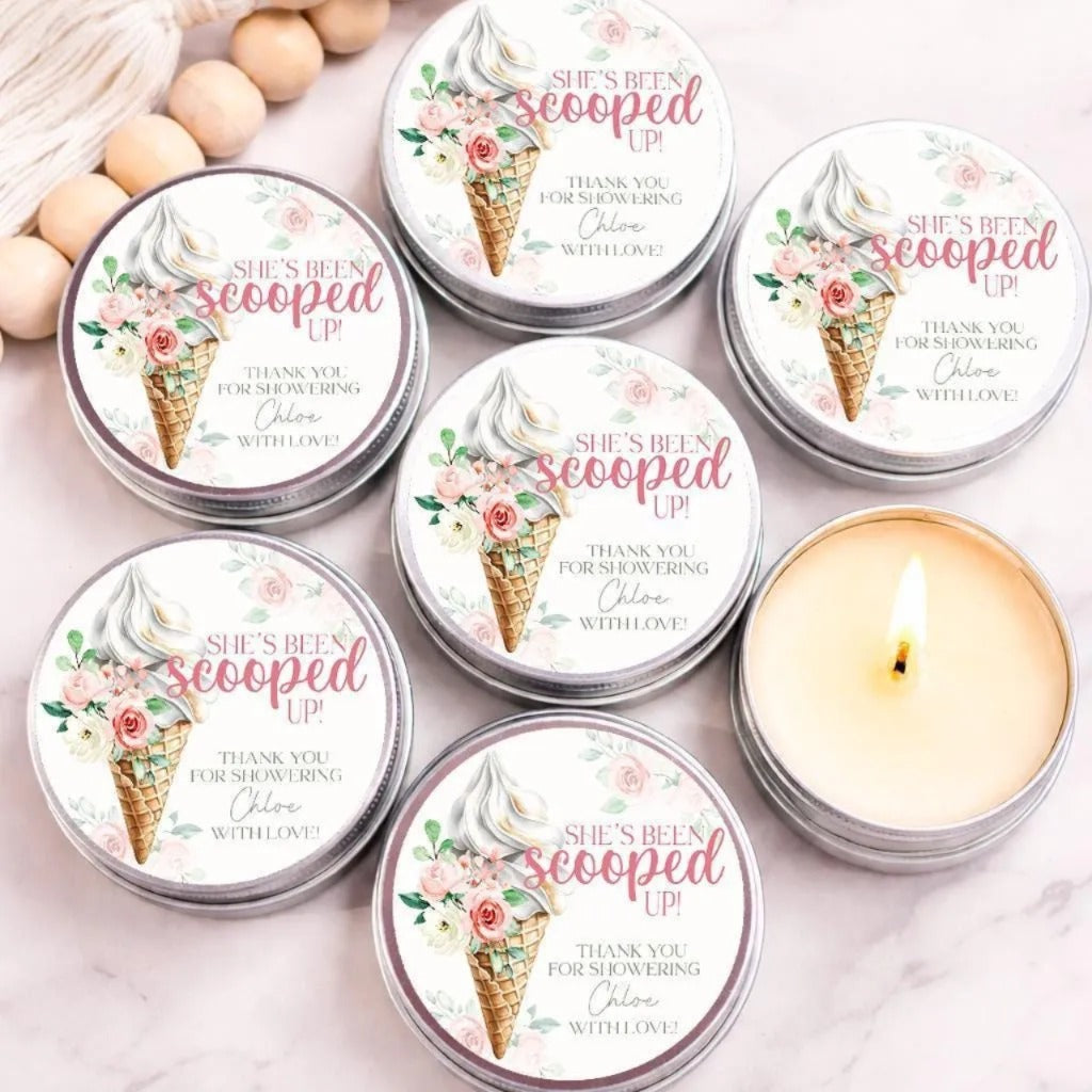 Personalized bridal shower candle favors displayed in silver tins on a light marble surface with a decorative wooden bead garland. Each candle features a watercolor ice cream cone topped with soft-serve ice cream, pink roses, and the phrase “She’s Been Scooped Up!” One candle is shown lit, highlighting the creamy wax. Perfect for ice cream-themed bridal showers, dessert-inspired celebrations, and bride-to-be party favors.