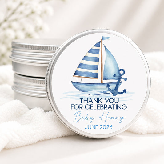 Personalized nautical baby shower candle favor in a silver tin featuring a watercolor sailboat with blue striped sails, a navy anchor, and custom text reading "Thank You for Celebrating Baby Henry" with the event date. Displayed on a soft white blanket with a neutral background, perfect for a nautical, sailing, or coastal-themed baby shower.