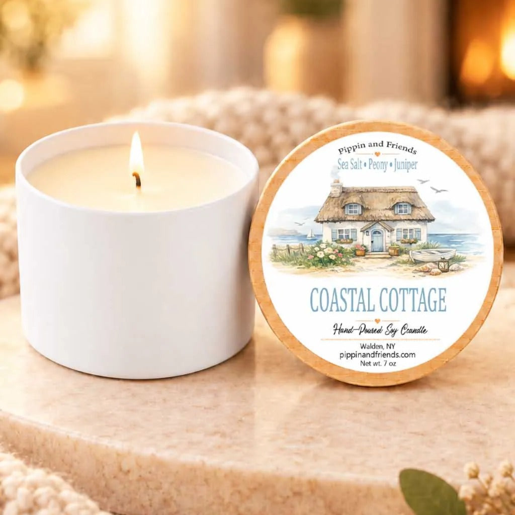 a white candle labeled "Coastal Cottage" next to a white candle holder, both sitting on a wooden surface.