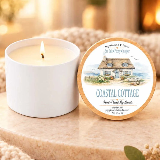 a white candle labeled "Coastal Cottage" next to a white candle holder, both sitting on a wooden surface.