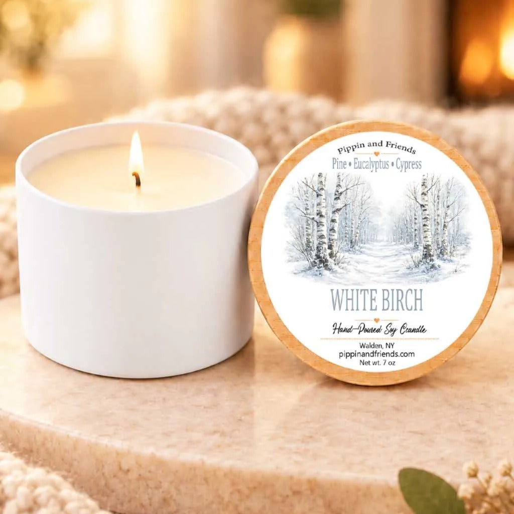 a white candle with a label that reads "White Birch" and "Pippin and Friends" next to it. The candle is placed on a wooden surface, and there is a blurred background that includes a fireplace.