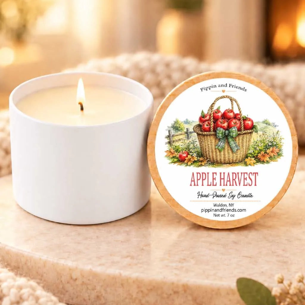 a white candle with a label that reads "Apple Harvest" and "Pippin and Friends", placed on a wooden surface next to a white candle holder.