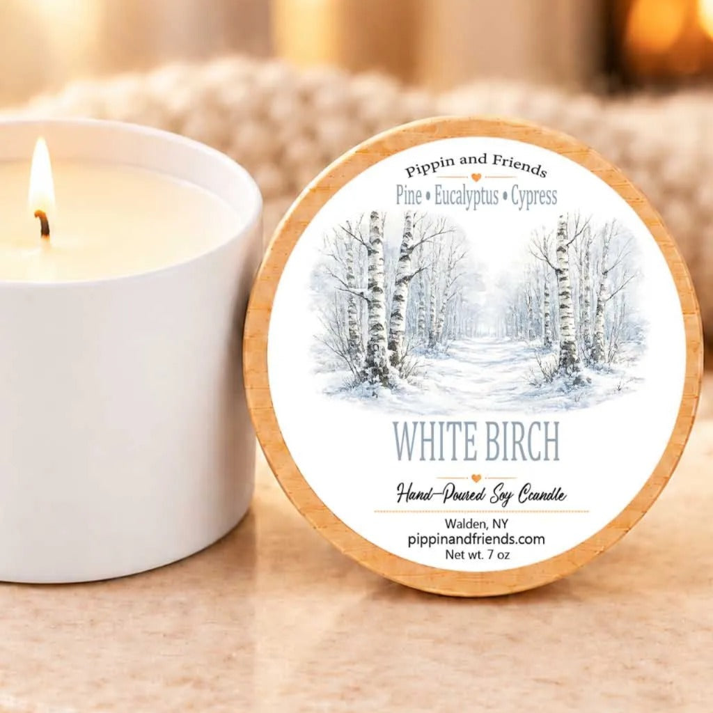 A white candle labeled "White Birch" sits next to a white candle, both placed on a wooden surface.