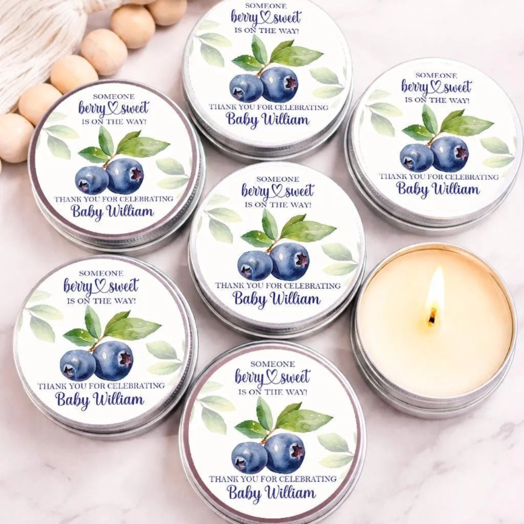Group of personalized berry sweet baby shower candle favors displayed in silver tins on a marble surface. Each candle features a watercolor strawberry design with delicate greenery and the phrase "Someone Berry Sweet Is On The Way" above custom text reading "Thank You for Celebrating Baby Lucy." One open candle reveals creamy wax, while decorative wooden beads and a tassel accent the background.