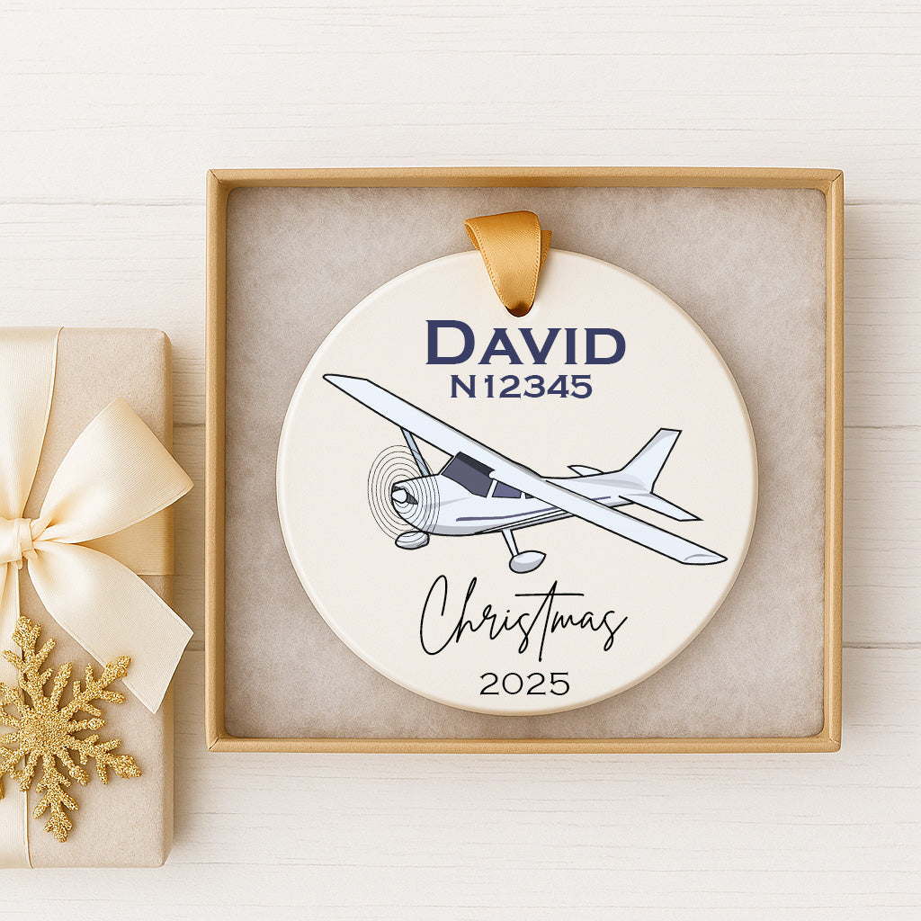 A white ceramic ornament with a blue airplane illustration and the name "DAVID" and the year "2025" printed on it, displayed in a gold frame.