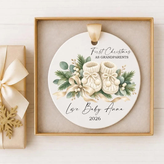 A white ceramic ornament with a gold frame, featuring a pair of baby booties and greenery, placed inside a gold box.