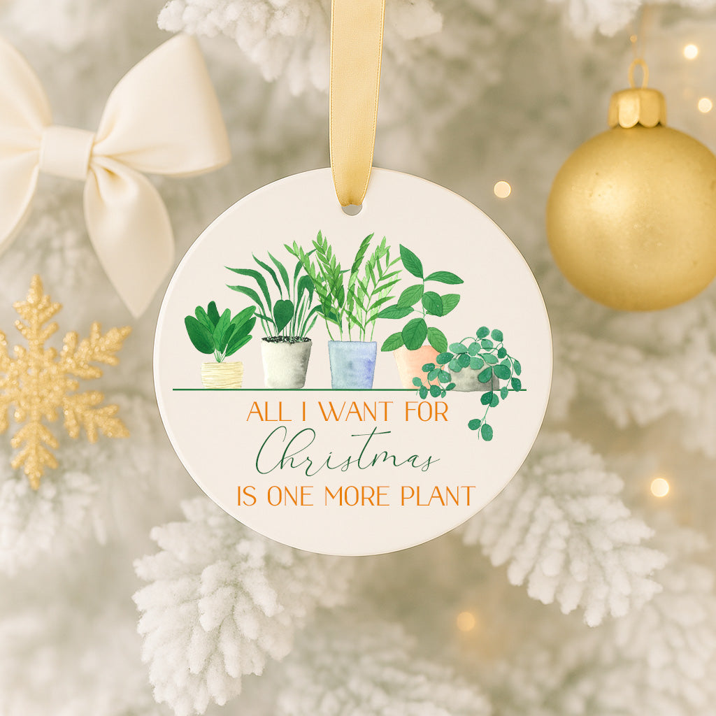 A white ornament with a gold ribbon hangs on a Christmas tree, featuring a potted plant and the text "All I want for Christmas is one more plant".