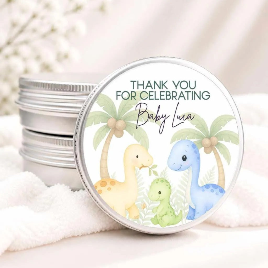 A round tin lid with a label featuring illustrations of two dinosaurs, one yellow and one blue, along with a message that says "Thank you for celebrating Baby Luca".