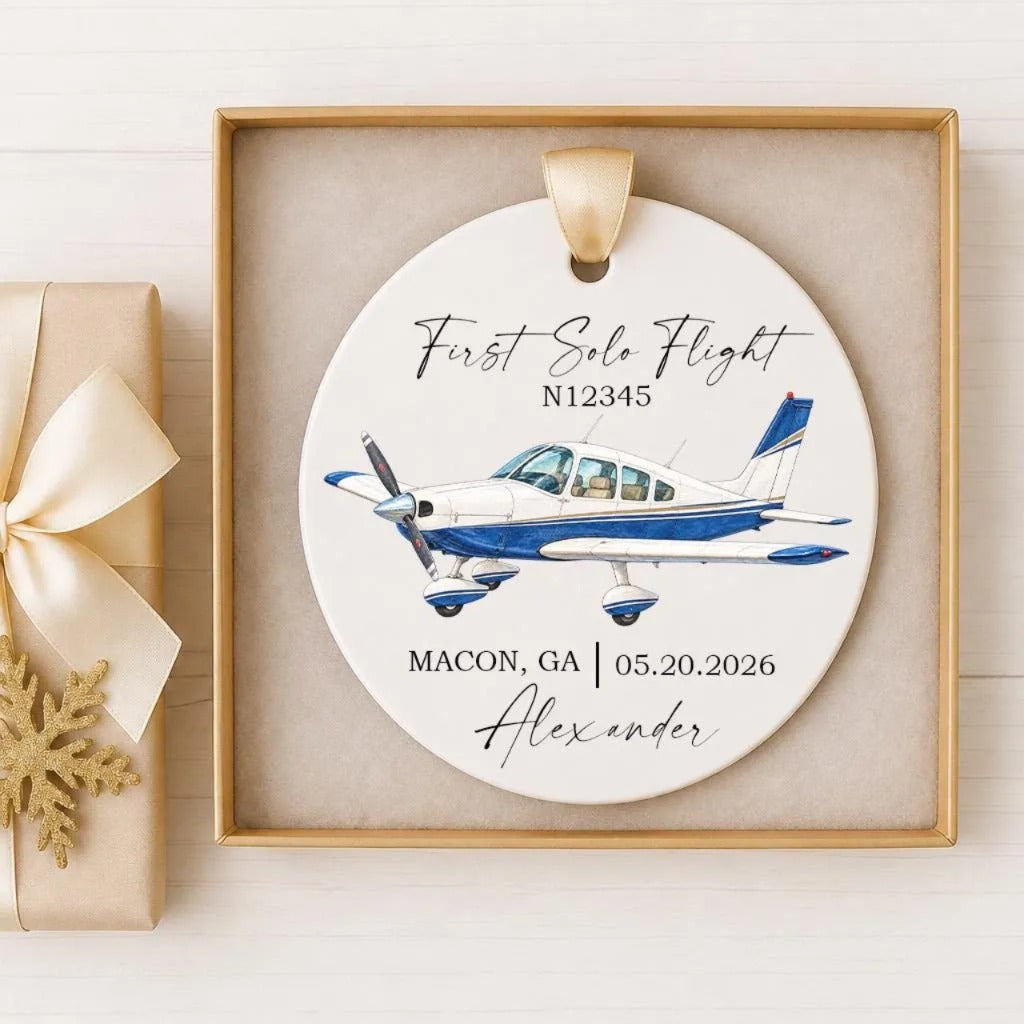A white ceramic ornament with a blue and white airplane illustration, along with the text "First Solo Flight N12345" and the location "Macon, GA 05.20.2026 Alexander".