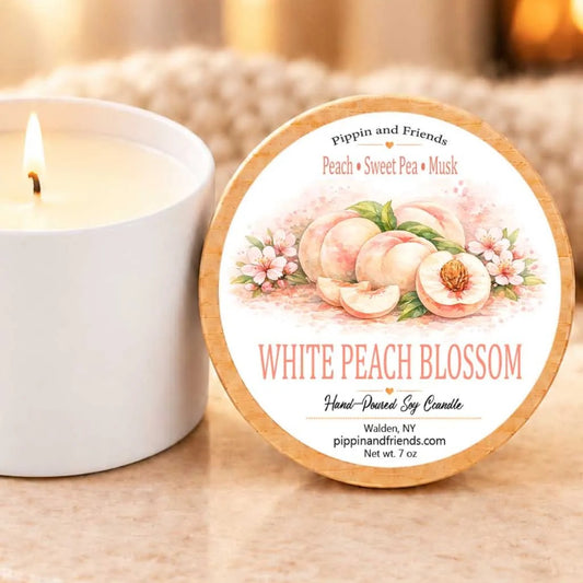 A white candle with a label that reads "White Peach Blossom" next to a lit candle on a table.