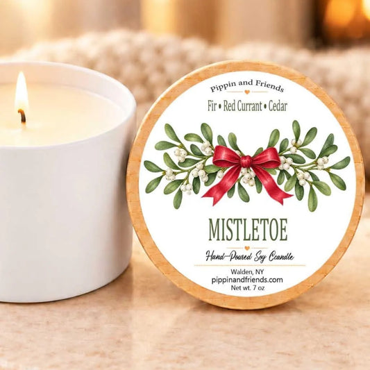 A white candle with a label that reads "Mistletoe Hand-poured Soy Candle" next to it, with a blurred background that appears to be a cozy, warm setting.
