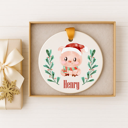A white ceramic ornament with a picture of a pig wearing a Santa hat, surrounded by greenery and berries, is displayed in a gold box.