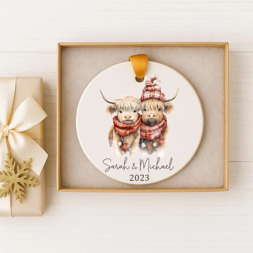 A ceramic ornament featuring two cows wearing winter clothing, with the text "Sarah & Michael" and the year "2023" printed on it. The ornament is displayed in a gold box, and there is a gift box with a gold ribbon next to it.