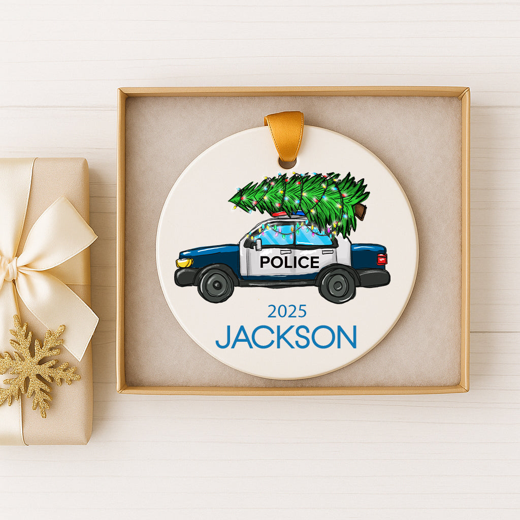 A white ornament with a blue police car and a Christmas tree on top, placed inside a gold box.