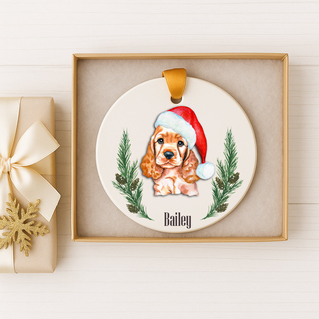A ceramic ornament featuring a brown and white dog wearing a Santa hat, surrounded by pine branches and a snowflake. The ornament is displayed in a gold box, and there is a gift box with a gold ribbon next to it.