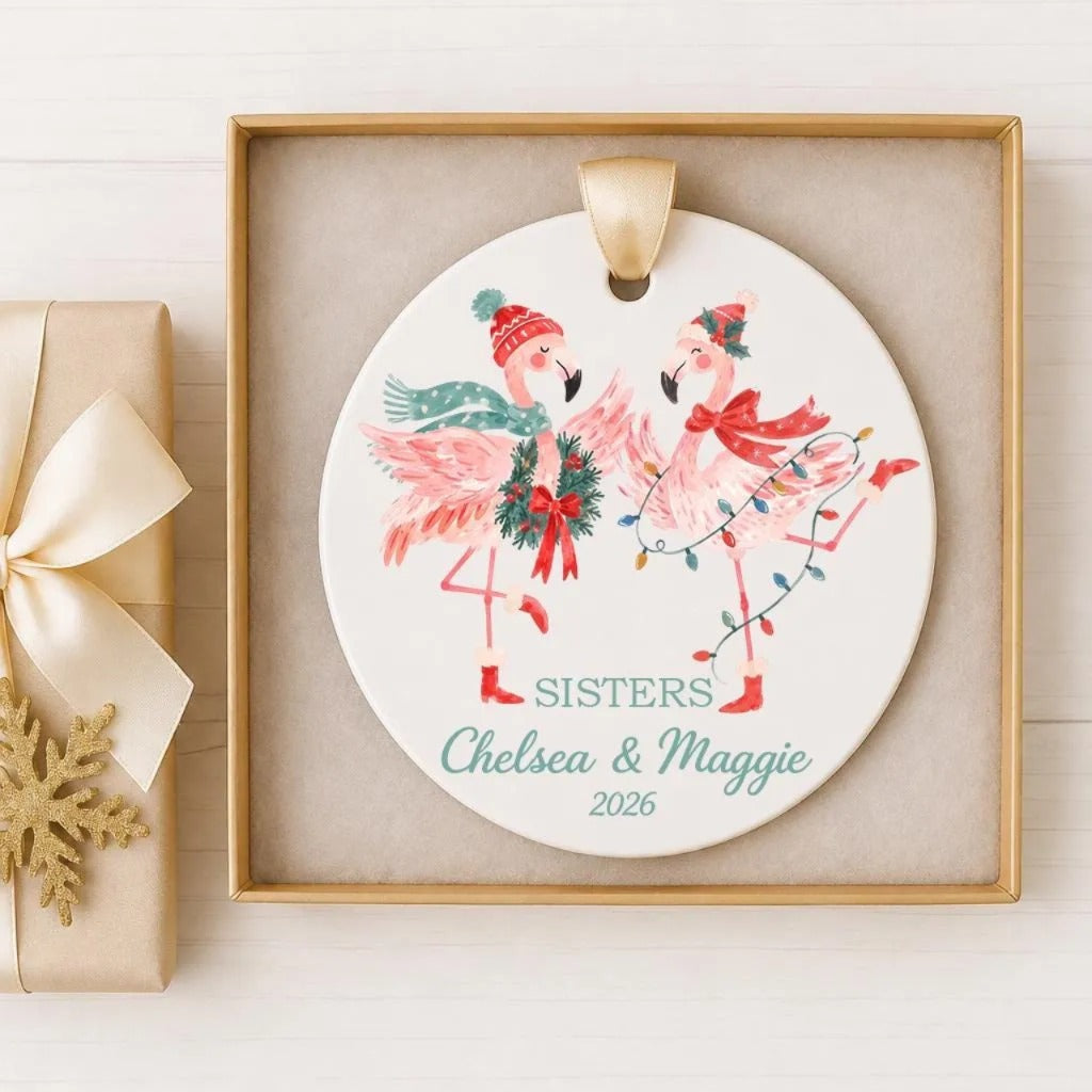 A decorative ornament featuring two pink flamingos, one wearing a red hat and the other a green hat, dancing together. The ornament is displayed in a gold frame, and there is a gift box with a ribbon on the side.