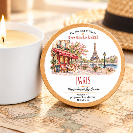 A round scented soy candle tin with a Paris-themed illustration label sits on a vintage map next to a lit white candle. The label reads 'Pippin and Friends' and 'PARIS' with scent notes Rose, Magnolia, and Patchouli.