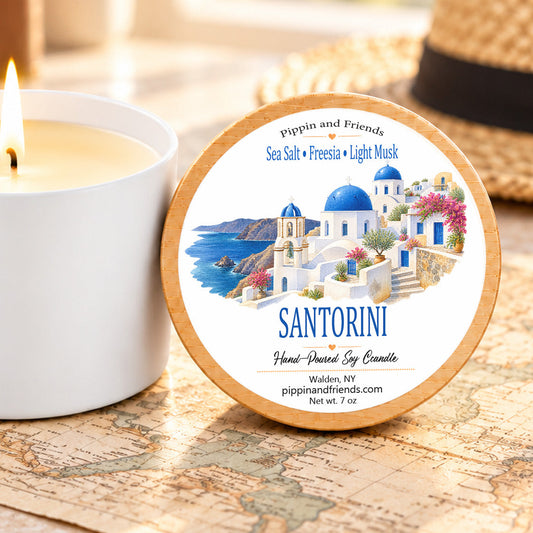 A round tin of Santorini hand-poured soy candle sits on a vintage map next to a lit white candle. The tin features a colorful illustration of a Greek island with blue domes and white buildings, and the brand name 'Pippin and Friends' is visible on the label.