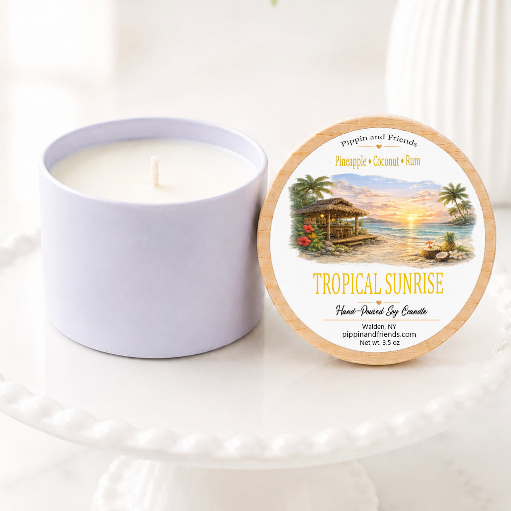A soy jar candle next to its round lid, which features a tropical sunset illustration and the text 'TROPICAL SUNRISE' and 'pippin and friends'. The items are styled on a white ceramic pedestal with a soft-focus background of green leaves and white flowers.