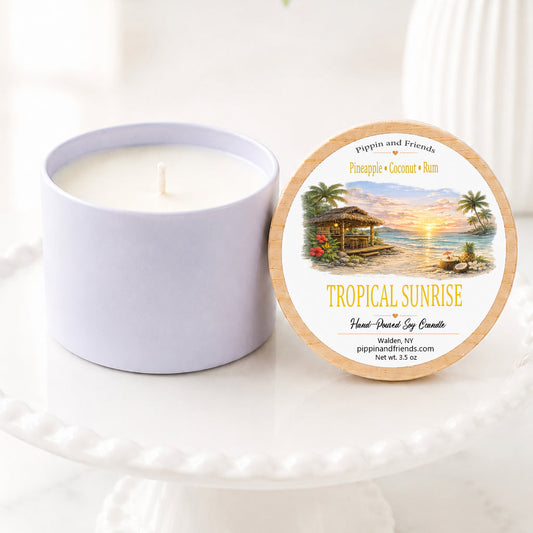 A soy jar candle next to its round lid, which features a tropical sunset illustration and the text 'TROPICAL SUNRISE' and 'pippin and friends'. The items are styled on a white ceramic pedestal with a soft-focus background of green leaves and white flowers.