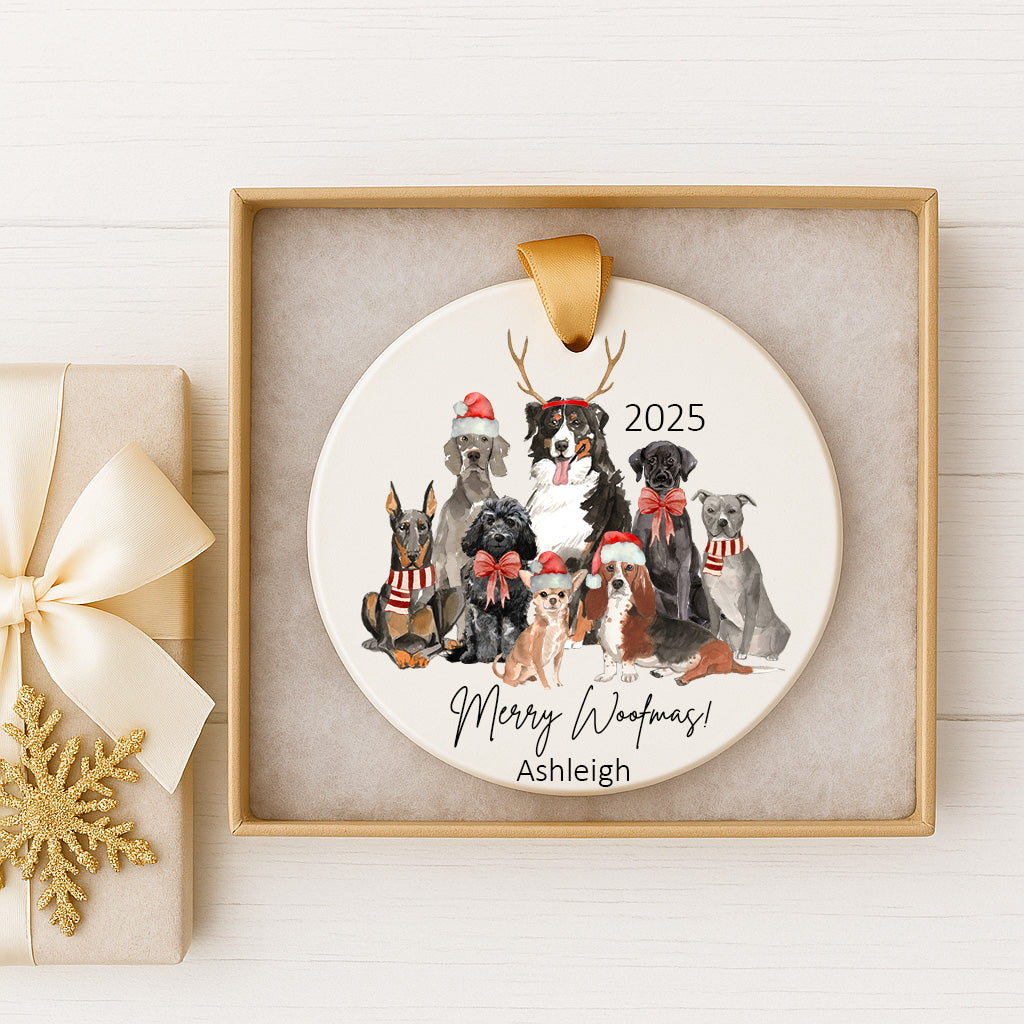 A white ceramic ornament with a festive design of dogs wearing Santa hats and scarves, along with the text "Merry Woofmas!" and "Ashleigh" printed on it. The ornament is displayed in a gold frame, and there is a gift box with a gold ribbon next to it.