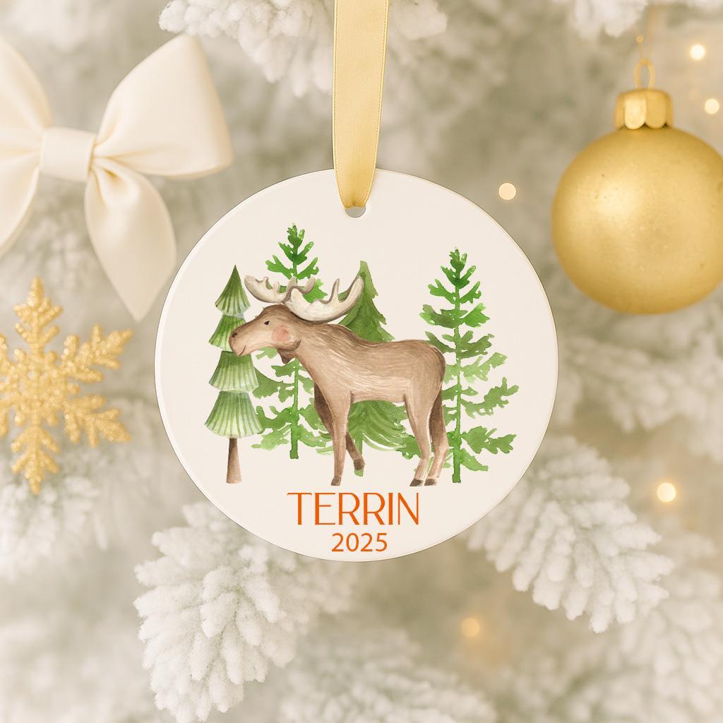 A white ornament with a brown moose and green trees hangs on a Christmas tree, surrounded by other ornaments and a gold ornament.