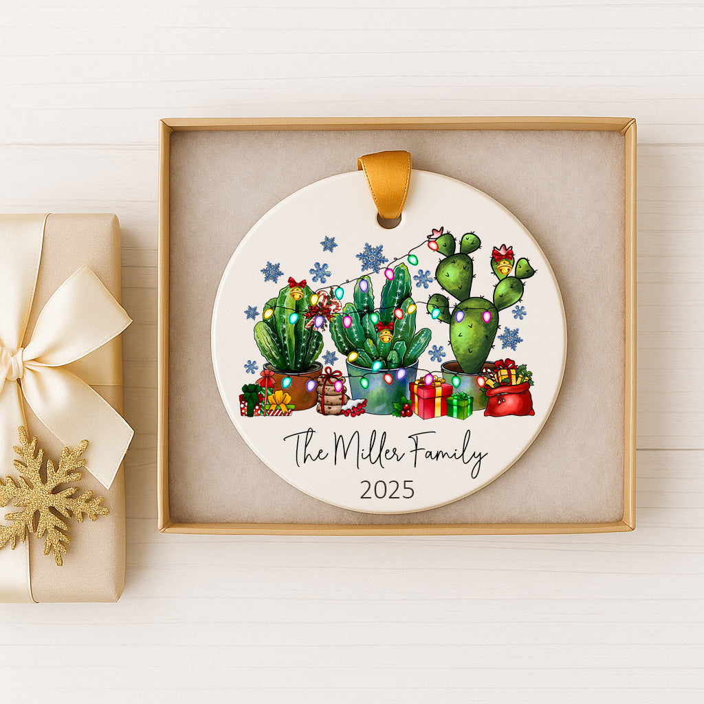 A white ceramic ornament with a festive design featuring cacti and Christmas lights, placed in a gold box.