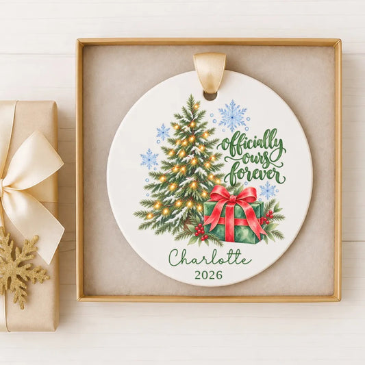 A white ceramic ornament with a Christmas tree and a gift box, placed in a gold frame on a white surface.