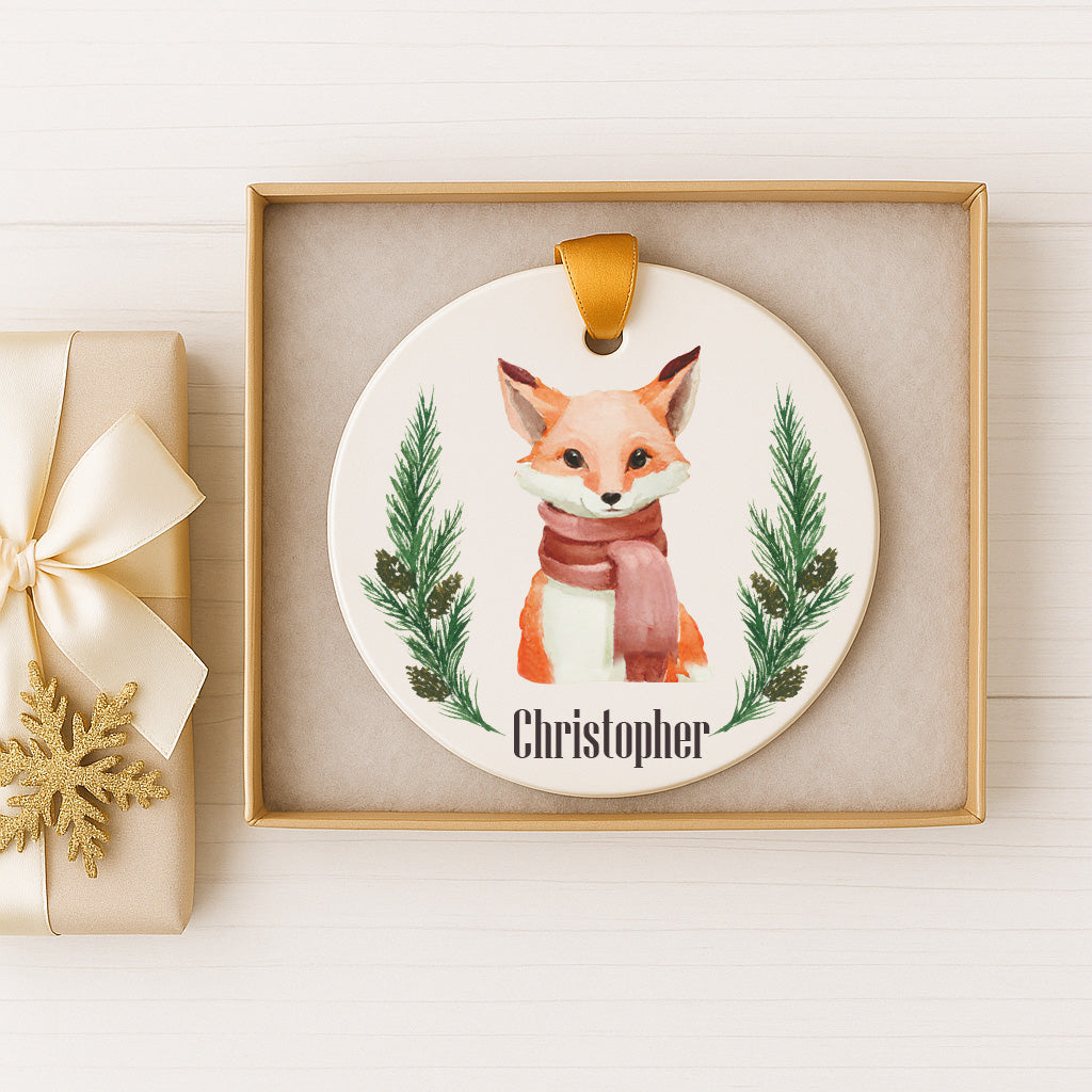 A ceramic ornament featuring a fox wearing a red scarf, surrounded by pine branches and a snowflake, is displayed in a box.