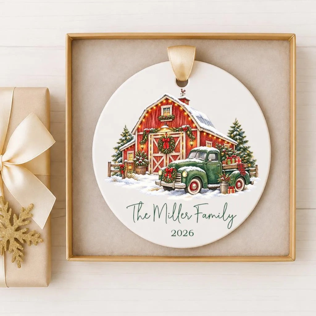 A white ceramic ornament with a red barn and green truck, surrounded by Christmas decorations, is displayed in a gold frame.