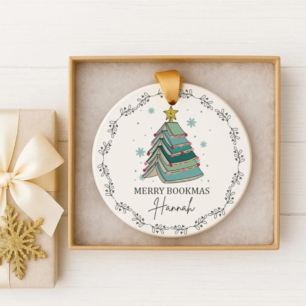 A white Christmas ornament with a tree design and the text "MERRY BOOKMAS Hannah" is displayed in a wooden box.