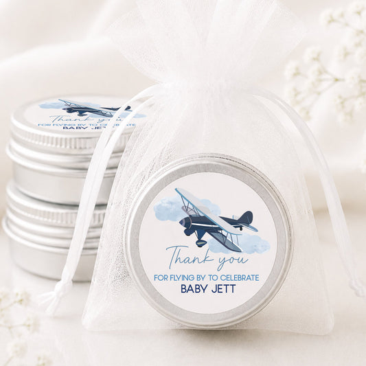 A small round silver tin wrapped in a sheer white organza drawstring bag, with a printed airplane label that reads 'Thank you FOR FLYING BY TO CELEBRATE BABY JETT.' Two more matching tins are stacked behind it on a soft white fabric background with delicate floral props.