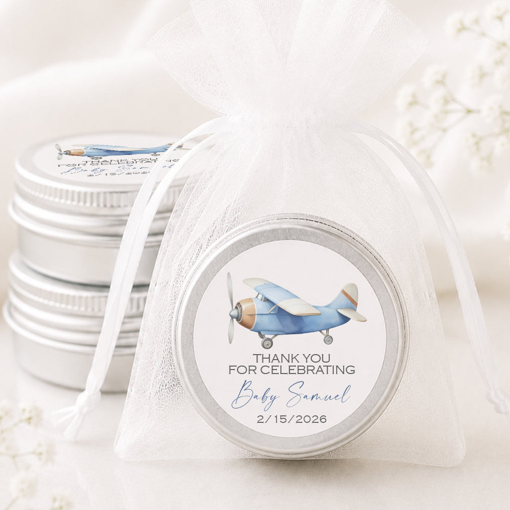 A small round silver tin wrapped in a sheer white organza bag with a printed airplane-themed favor label reading 'Thank you for celebrating Baby Samuel 2/15/2026.' Additional matching favor tins are stacked in the background on a soft white surface with delicate floral props.