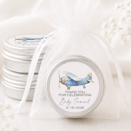 A small round silver tin wrapped in a sheer white organza bag with a printed airplane-themed favor label reading 'Thank you for celebrating Baby Samuel 2/15/2026.' Additional matching favor tins are stacked in the background on a soft white surface with delicate floral props.