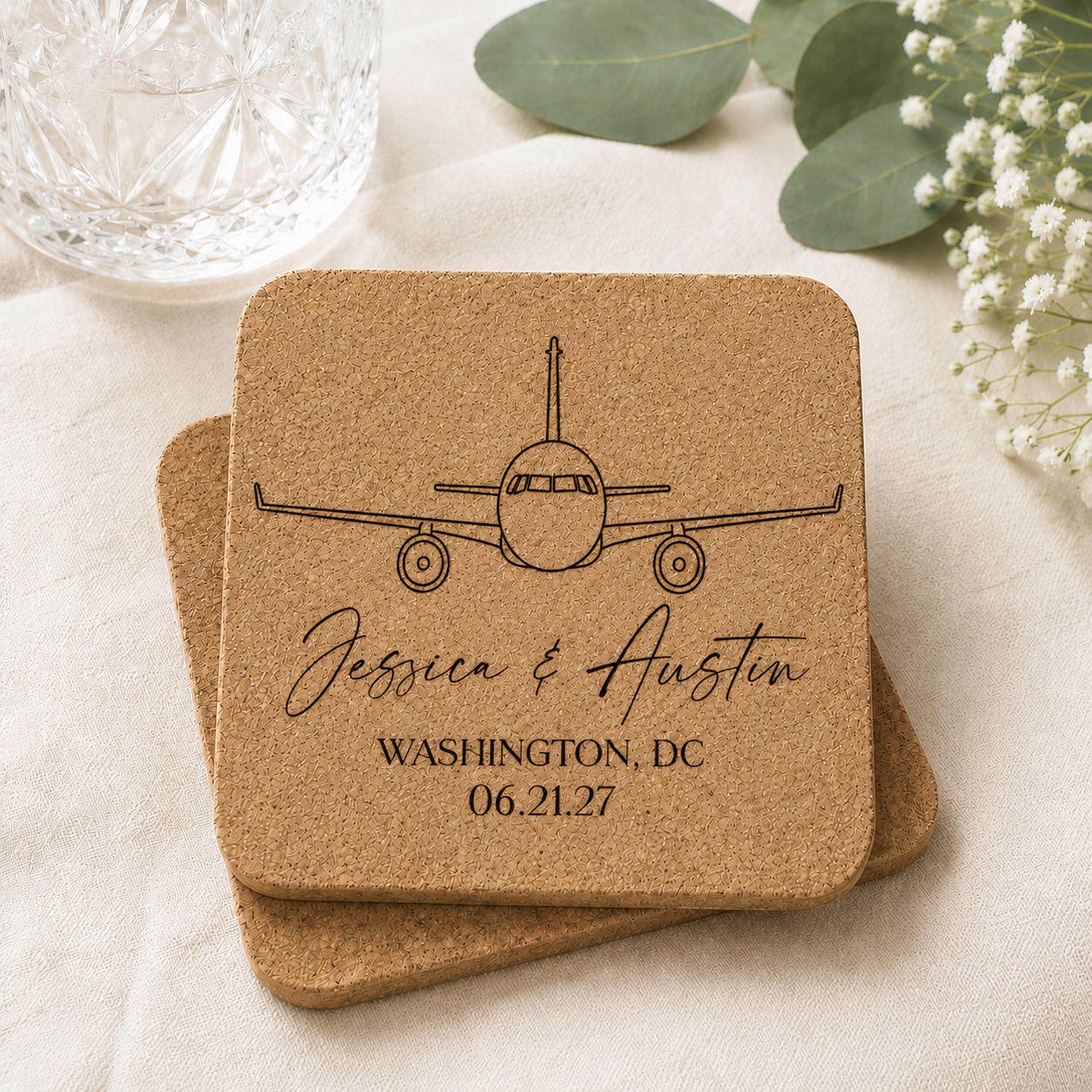 Personalized cork coaster featuring a laser engraved airplane design with custom names, wedding location, and wedding date. Natural cork coasters are displayed stacked on a soft neutral background with greenery and crystal glassware.
