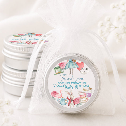 A round silver tin wrapped in a sheer white organza bag, with a printed round label that reads 'Thank you for celebrating Violet's 1st Birthday With Us' and features pastel illustrations of a girl in a blue dress, pink roses, and a teapot. More matching silver tins are stacked in the background on a soft white surface.