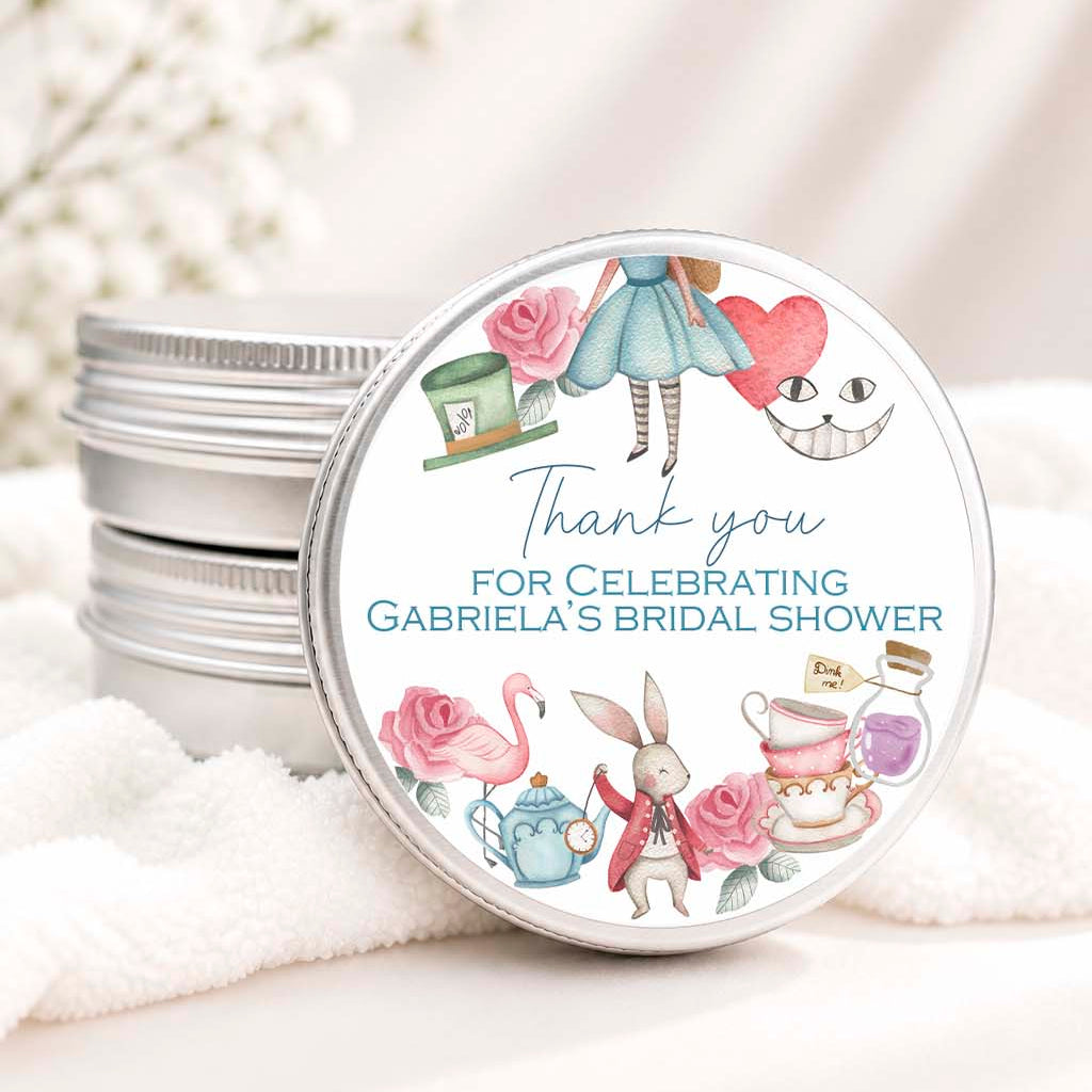A round silver tin lid with a personalized bridal shower thank you label, shown in front of stacked matching silver tins on a soft white fabric surface with blurred white flowers in the background.