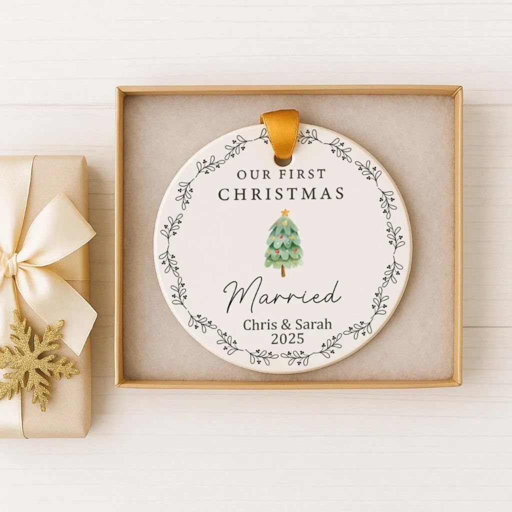 A white ceramic ornament with a gold ribbon, placed in a gold box. The ornament features a Christmas tree and the text "Our first Christmas Married Chris & Sarah 2015".