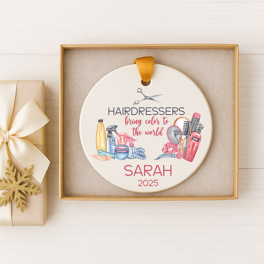 A decorative ornament in a gold box, featuring illustrations of hair styling tools and text that reads "HAIRDRESSERS bring color to the world" and "SARAH 2025".