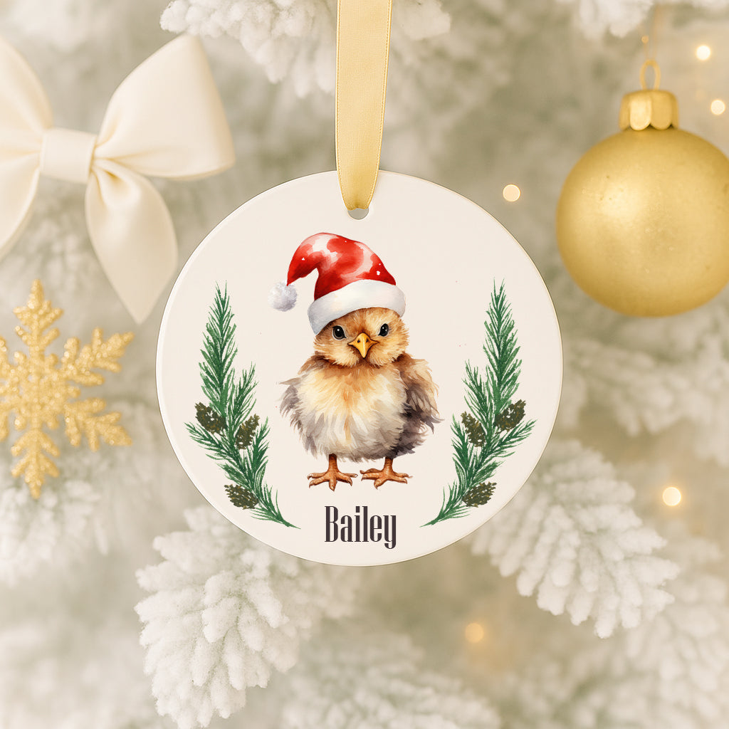 A white ornament with a brown and white chicken wearing a Santa hat, surrounded by pine branches and pine cones, is hanging on a Christmas tree.