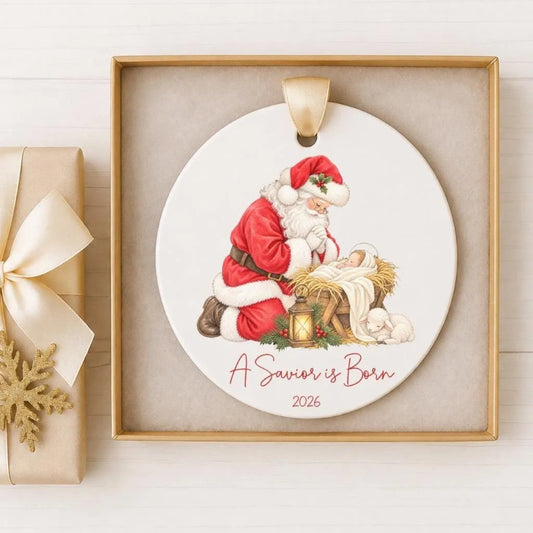 A ceramic ornament depicting Santa Claus and a baby Jesus in a nativity scene, with the text "A Savior is Born" and the year "2026" below.
