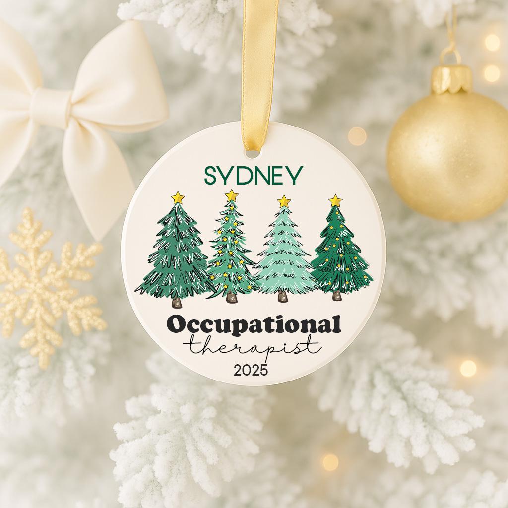 A white ornament with a gold ribbon hangs on a Christmas tree, featuring a design of four green Christmas trees with a star on top.