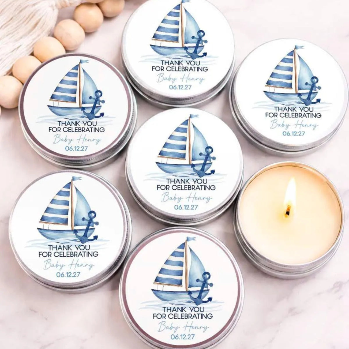 a collection of six tin candles with a nautical design, arranged on a marble surface. The candles are labeled with "Thank you for celebrating Baby Henry" and "06.12.27".