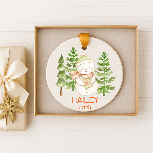 A white ornament with a snowman and pine trees, displayed in a box with a gold ribbon.