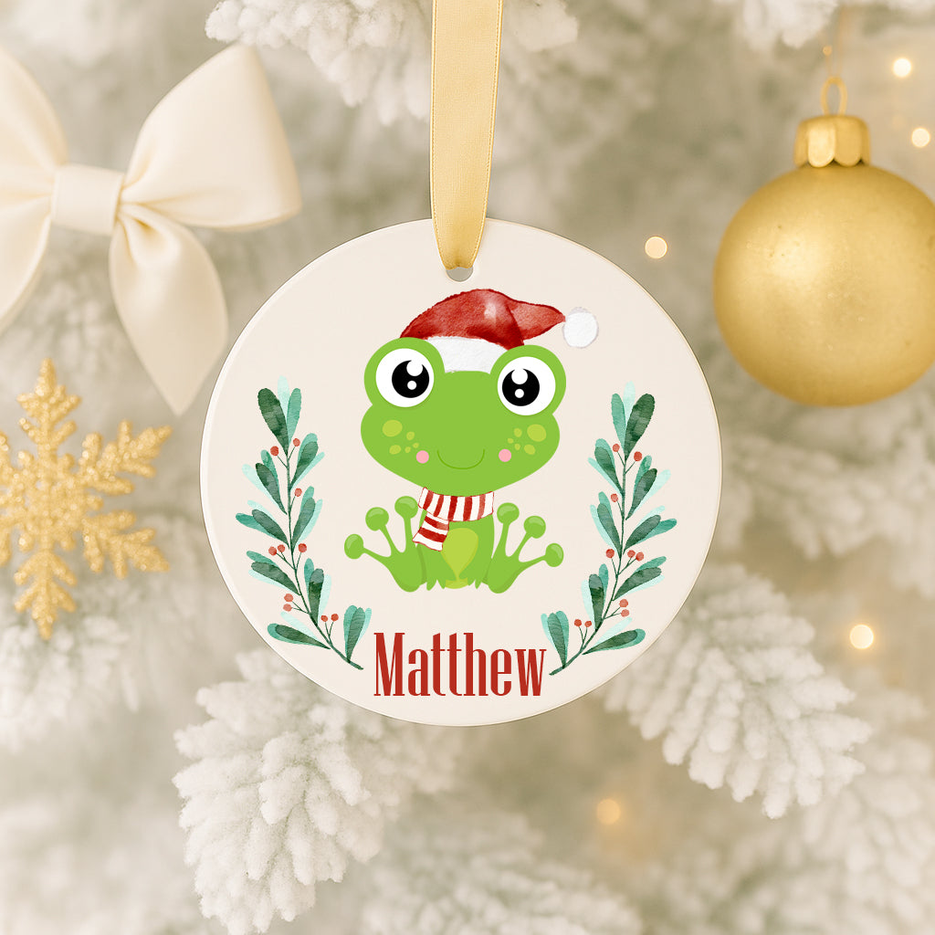 A white ornament with a green frog wearing a Santa hat, surrounded by a wreath of greenery and the name Matthew.