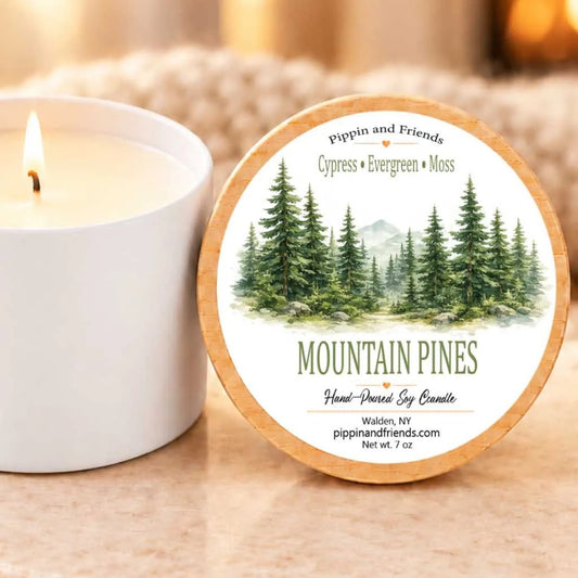 a white candle with a label that reads "Mountain Pines" and "Pippin and Friends" on a wooden holder. The candle is placed next to a blurred background, which appears to be a cozy indoor setting with a fireplace.