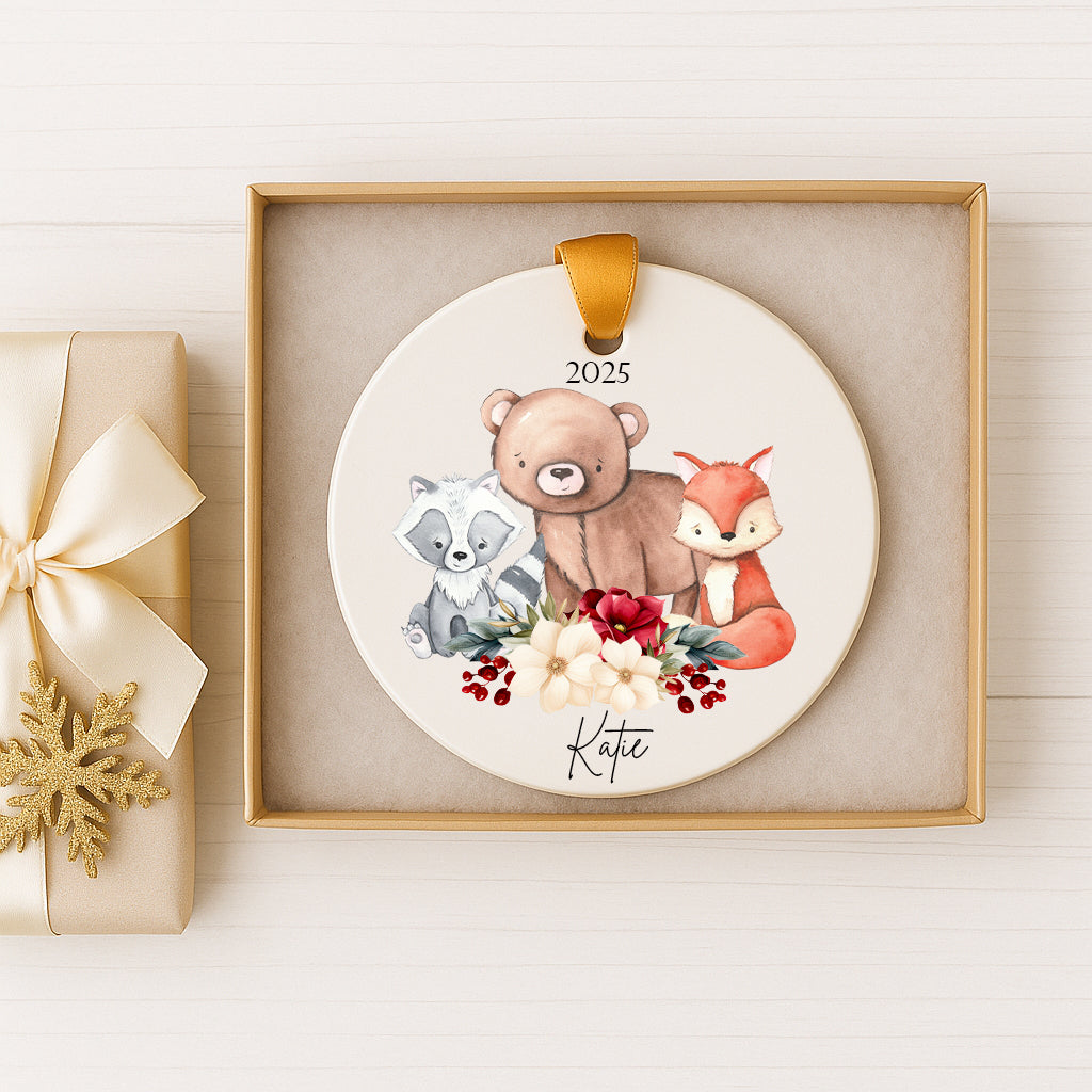 A decorative ornament featuring a bear, raccoon, and fox surrounded by flowers and berries, with the name "Katie" written on it. The ornament is displayed in a gold frame, and there is a gift box with a bow next to it.