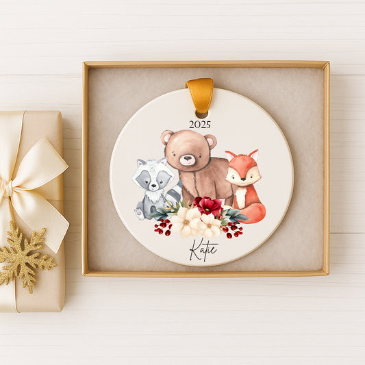 A decorative ornament featuring a bear, raccoon, and fox surrounded by flowers and berries, with the name "Katie" written on it. The ornament is displayed in a gold frame, and there is a gift box with a bow next to it.