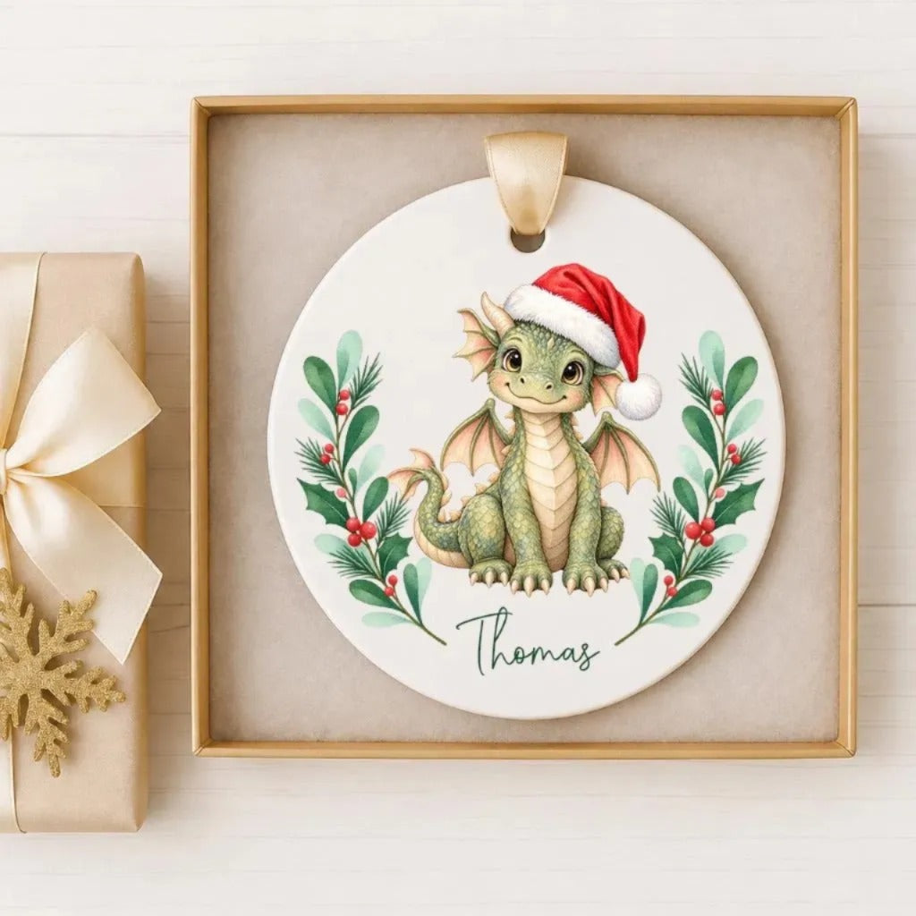 A ceramic ornament featuring a cartoon dragon wearing a Santa hat, surrounded by holly leaves and berries, with the name "Thomas" written below the dragon.