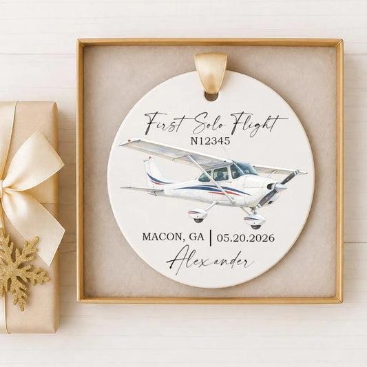 A white ceramic ornament with a gold ribbon, featuring an illustration of a small airplane and the text "First Solo Flight N12345" and "Macon, GA 05/20/2026 Alexander".